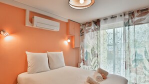 Uma cama bem arrumada com almofadas, um ar condicionado fixado na parede e uma janela com cortina estampada com folhas.