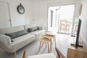 A modern living room with a sofa, a small table, a TV, and a balcony with outdoor furniture. - Clos des Sables B13 - Two Bedroom Apartment, Sleeps 4 (Les Herbiers)