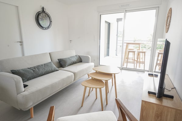 A modern living room with a sofa, a small table, a TV, and a balcony with outdoor furniture.