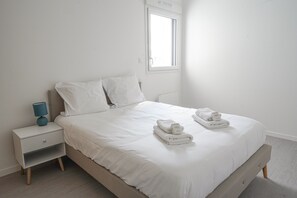 A neatly made bed with white linens and two towels, a bedside table with a lamp, and a window providing natural light.