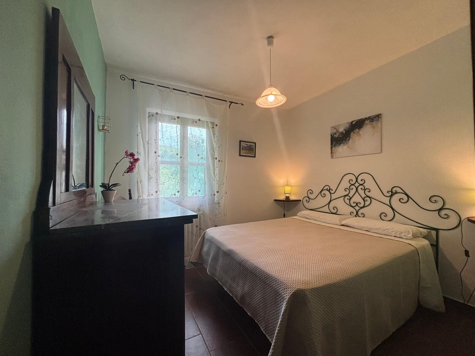 Una camera da letto con un letto, una lampada da comodino, una finestra con tende, un comò e uno specchio.
