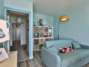 A room with a blue sofa, a bookshelf, a floor lamp, and a door leading to another room. - Reflets d'Océan - vue imprenable sur la mer (Les Sables d'Olonne)