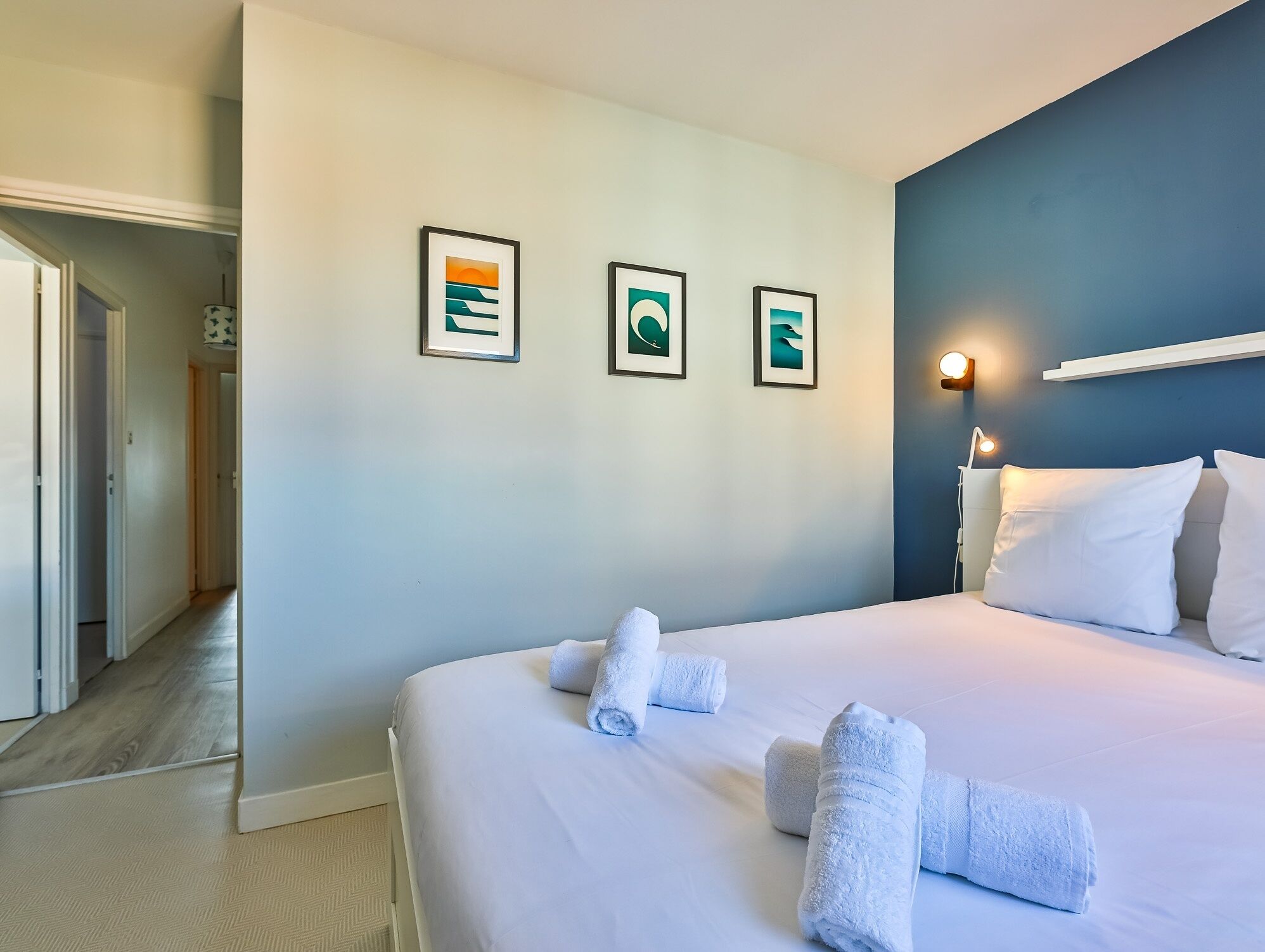 Une chambre d’hôtel avec un lit, des serviettes blanches, un couloir et trois tableaux encadrés accrochés au mur.