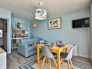 A modern living room with a sofa, dining table, chairs, a TV, a world map, and a bookshelf. - Reflets d'Océan - vue imprenable sur la mer (Les Sables d'Olonne)