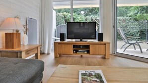 Una sala de estar moderna con un televisor de pantalla plana sobre un pedestal de madera, un sofá, una mesa de centro con una revista y vistas a un jardín a través de amplios ventanales.