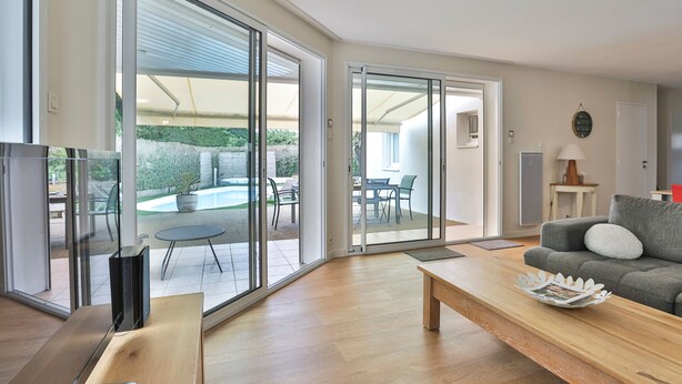 Una sala de estar moderna con un sofá, una mesa de centro y un televisor. Hay una puerta corrediza de vidrio que da a un patio exterior con piscina y sillas.