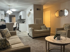 Ein Wohnzimmer mit einer Couch, Sesseln, einem Couchtisch und einer Küche im Hintergrund.