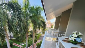 A balcony with a white table and chairs, a small potted plant, and a view of a well-maintained garden with palm trees and a pathway. - Ducassi Penthouses Rooft Pool (Punta Cana)