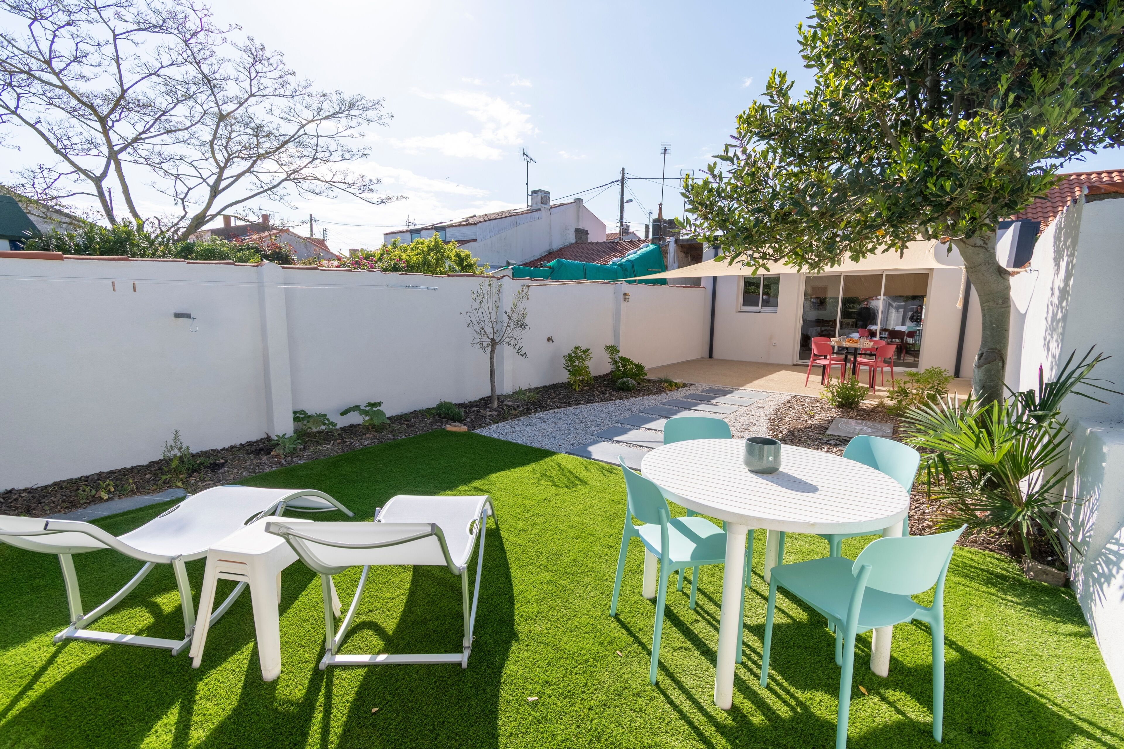 Un patio con una mesa y sillas blancas, un pequeño jardín con césped artificial y vista a un edificio cercano.