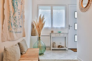 Una habitación con cama, un florero con plumas de pampa, una ventana con persianas, una puerta blanca y un espejo fijado a la pared.