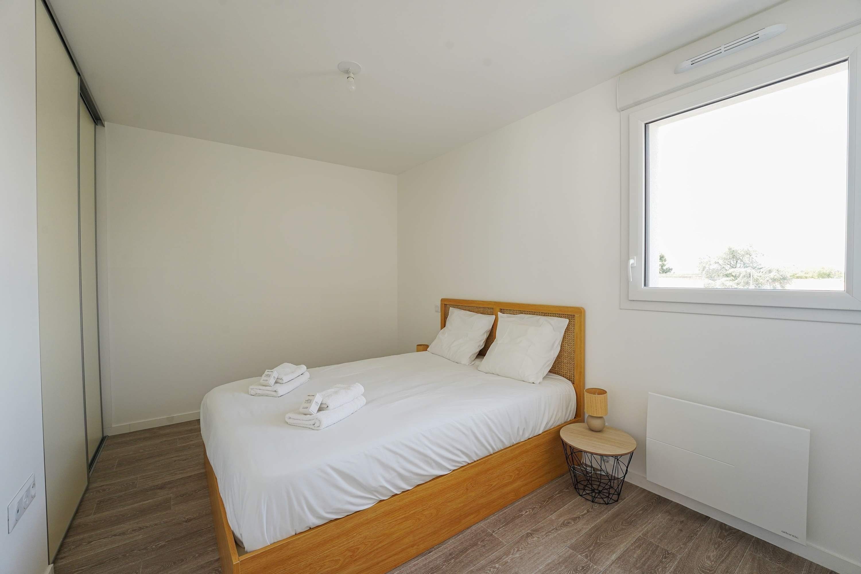 A modern bedroom with a wooden bed, white linens, a bedside table with a lamp, and a window with a view.