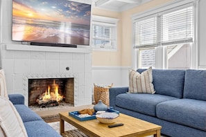 A living room with a fireplace, a blue sofa, a television, and a table with snacks. - Luxury Beach Block | Sauna + Cold Plunge + Hot Tub (Ventnor City)