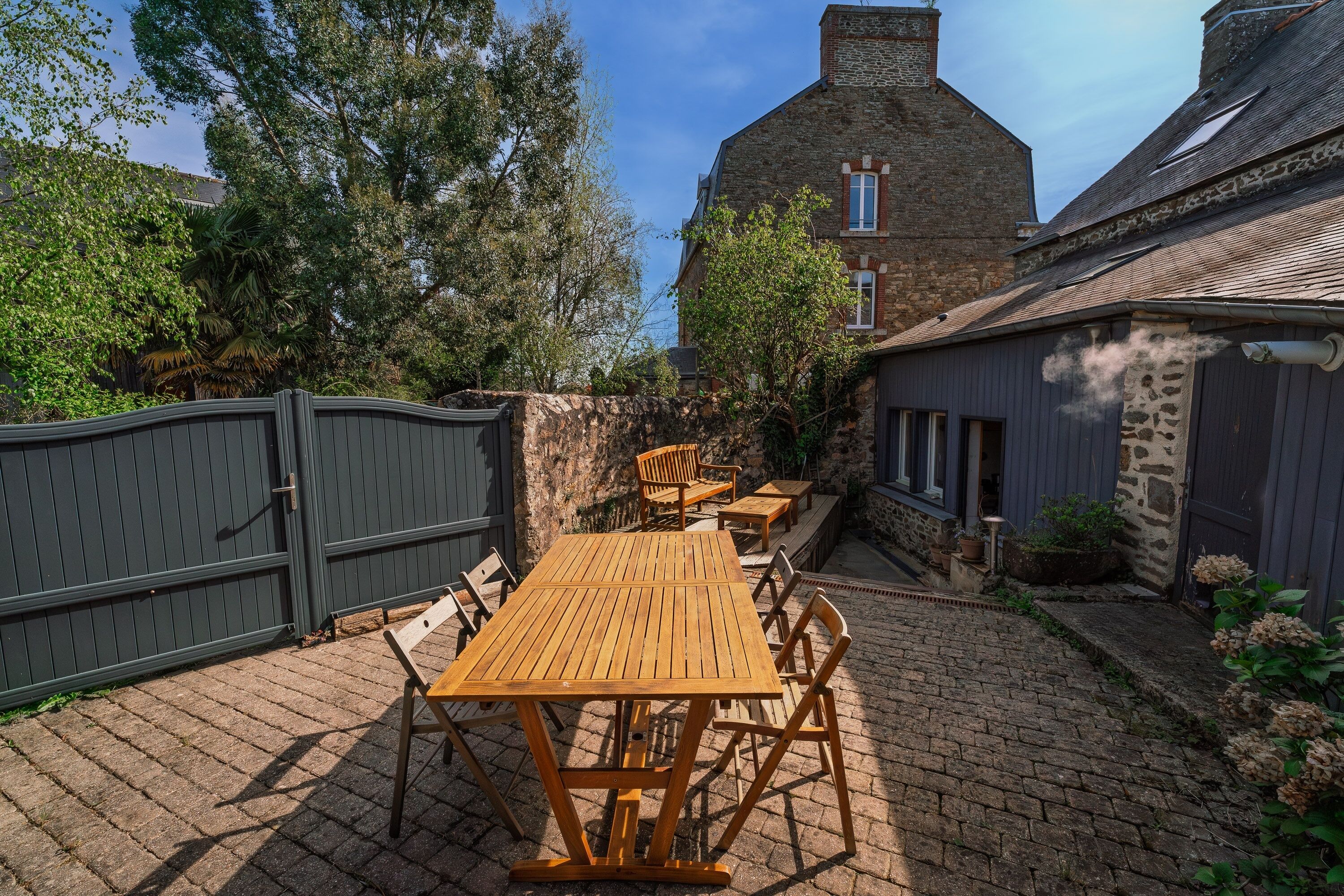 Une terrasse extérieure avec une table et des chaises en bois, un mur en pierre et, au fond, un grand bâtiment en briques.