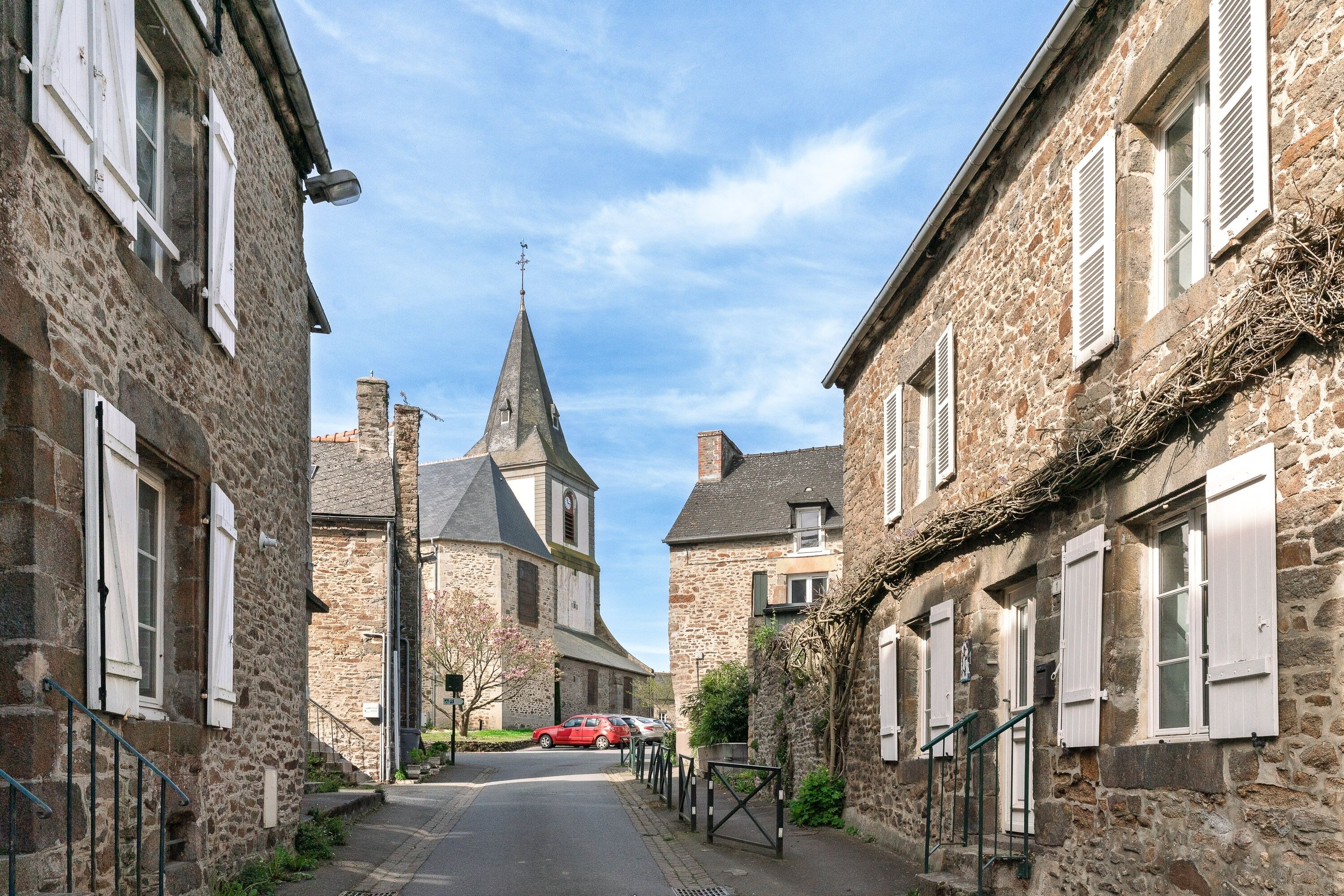 A narrow street lined with stone buildings, a church with a pointed steeple, and a red car parked on the side.