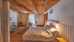 Una camera da letto con soffitto in legno, un letto con coperta bianca, una panca in legno, una sedia, un tavolino e una finestra con tende.