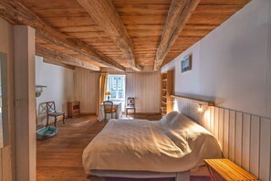 A bedroom with a wooden ceiling, a bed with a white cover, a wooden bench, a chair, a small table, and a window with curtains. - Ty Rance - Maison Avec Terrasse (Plouër-sur-Rance)