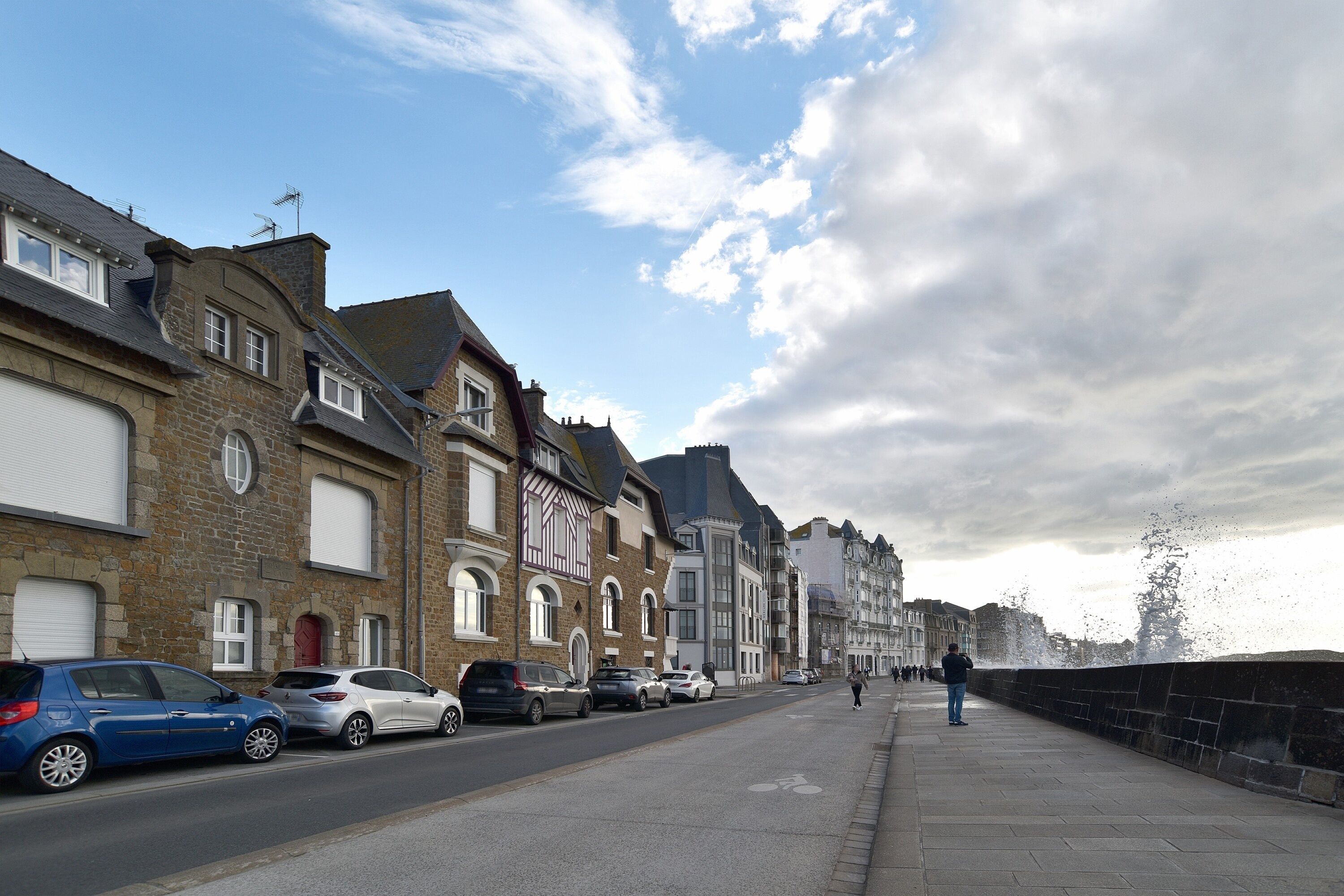 A street with parked cars, a pedestrian walkway, and buildings with traditional architecture.
