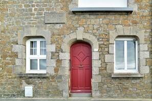 Ein Steinhaus mit roter Tür, zwei Fenstern und einer kleinen Tafel.