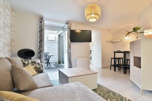 Ein modernes Wohnzimmer mit einer Couch, einem Couchtisch und einem an der Wand montierten Fernseher. Es gibt einen Essbereich mit Tisch und Stühlen sowie eine Küchenzeile mit einer Arbeitsplatte und Hockern.