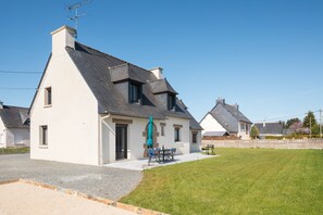 A two-story house with a sloped roof, a front door, and a small porch with a table and chairs. - La Fréhéloise - Jolie maison pour 9 avec jardin (Fréhel)