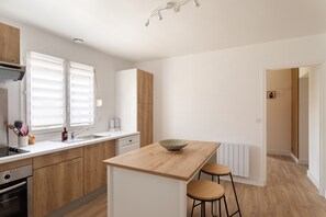 A modern kitchen with wooden cabinets, a central island, bar stools, and a view of the adjacent room. - La Fréhéloise - Jolie maison pour 9 avec jardin (Fréhel)