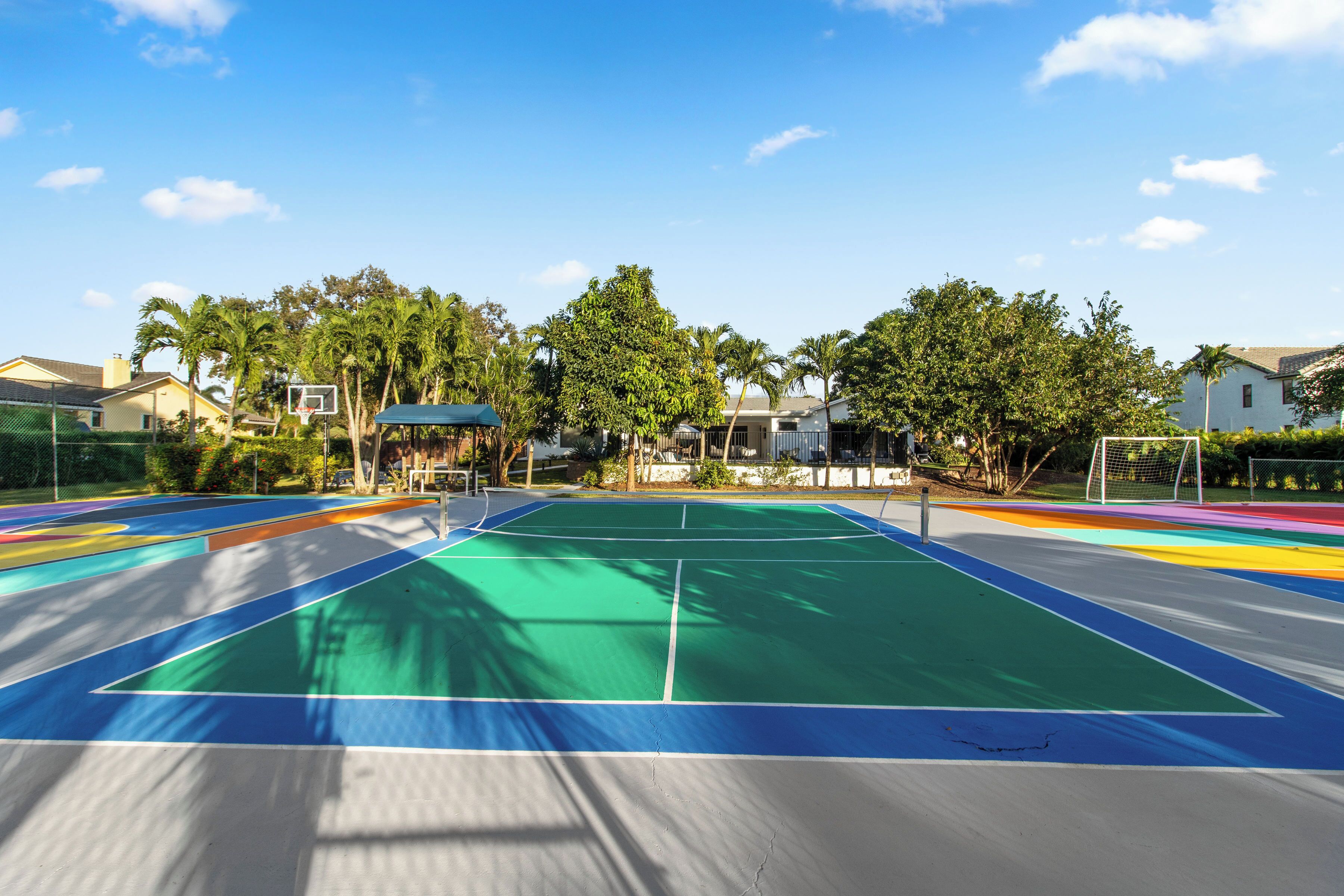 An outdoor basketball court with a green playing surface, surrounded by trees and houses.