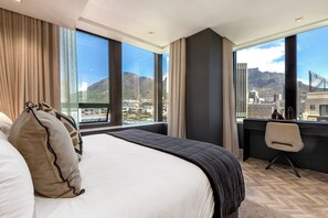 A hotel room with a large bed, a desk, and a view of a cityscape and mountains. - Foreshore Luxury Place (Cape Town City Centre)