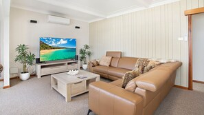 Een woonkamer met een flatscreen televisie, twee potplanten, een beige bank en een salontafel.