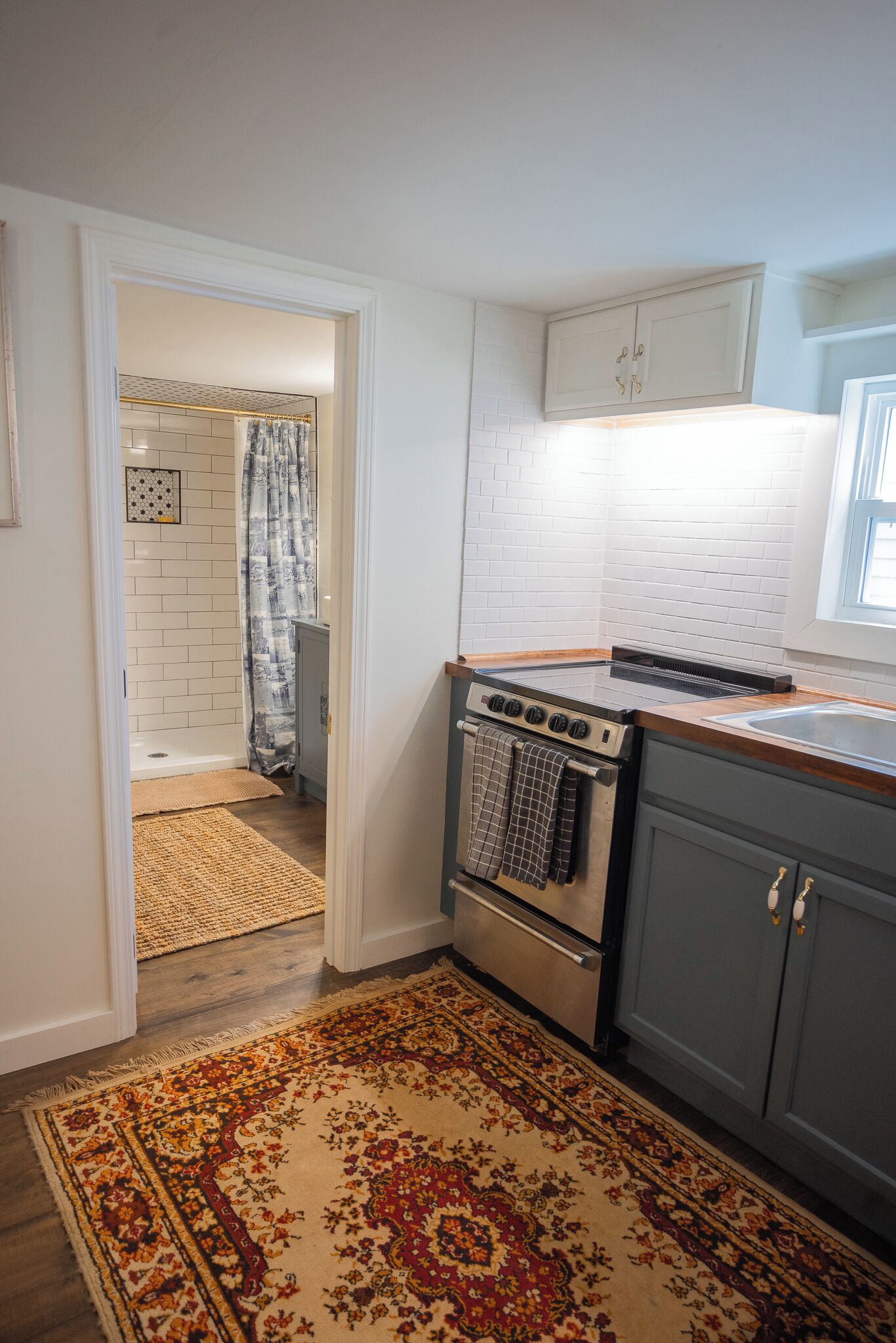 Une cuisine équipée d’une cuisinière, d’un four et de placards, donnant sur une salle de bain avec une douche et un tapis à motifs.