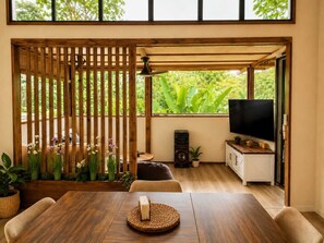 Une pièce comprenant une table à manger en bois, des chaises, un téléviseur, un haut-parleur et un balcon agrémenté de plantes.
