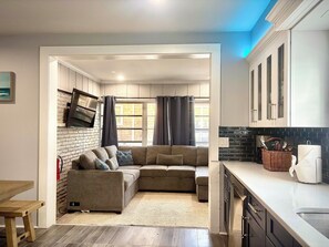 A modern living room with a sectional sofa, a flat-screen TV, and a kitchen with dark backsplash and white cabinets.