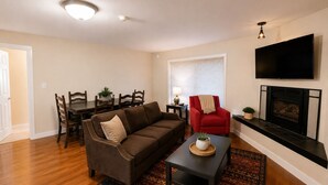 Un salon avec un canapé marron, un fauteuil rouge, une table à manger avec des chaises, une cheminée et un téléviseur fixé au mur.