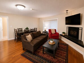 Ein Wohnzimmer mit braunem Sofa, einem roten Sessel, einem Esstisch mit Stühlen, einem Kamin und einem an der Wand montierten Fernseher.