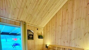 Ein Schlafzimmer mit Holzbalkendecke, einem Bett mit weißer Bettdecke, einem Nachttisch und einem Fenster mit Blick auf einen Außenbereich.