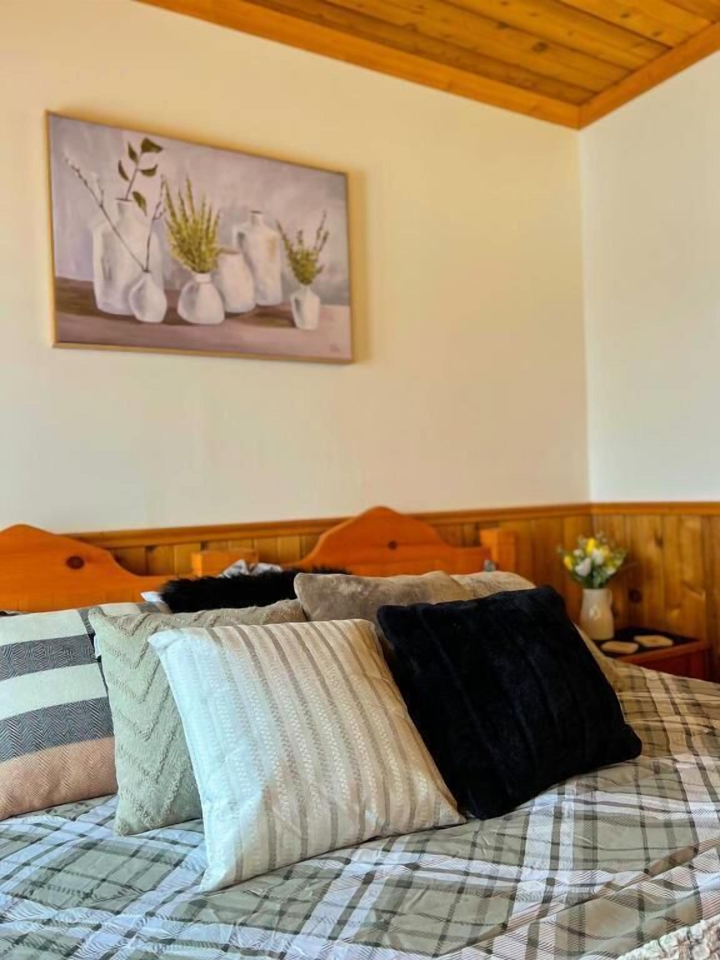 A bedroom with a bed, pillows, a headboard, a painting, and a nightstand with a vase.