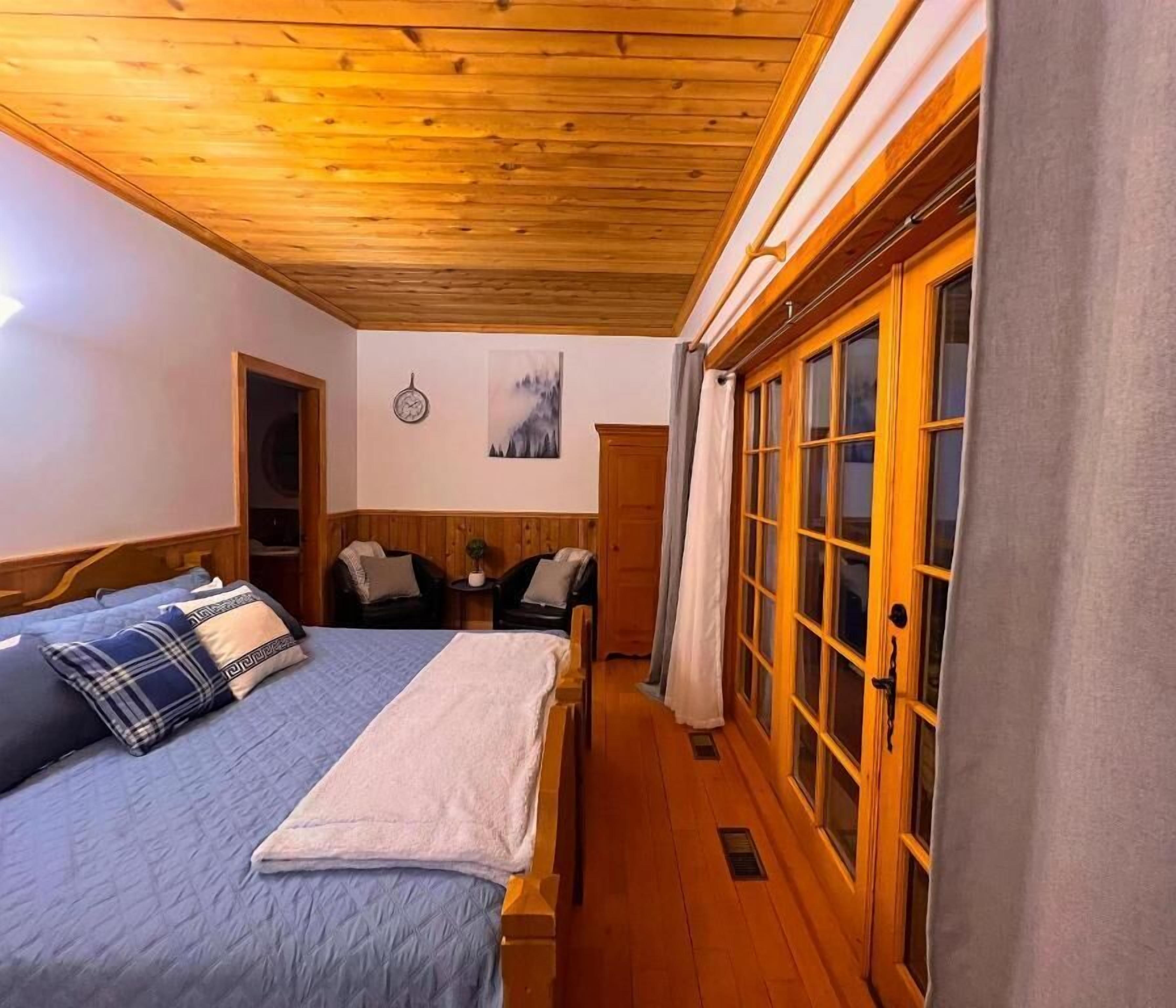 A bedroom with a wooden ceiling, a bed with a blue quilt, a wooden wardrobe, and a small table with chairs.