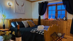 Un cuarto acogedor con cama, cabecera de madera, ventana con cortinas, una mesita con una botella y un reloj en la pared.