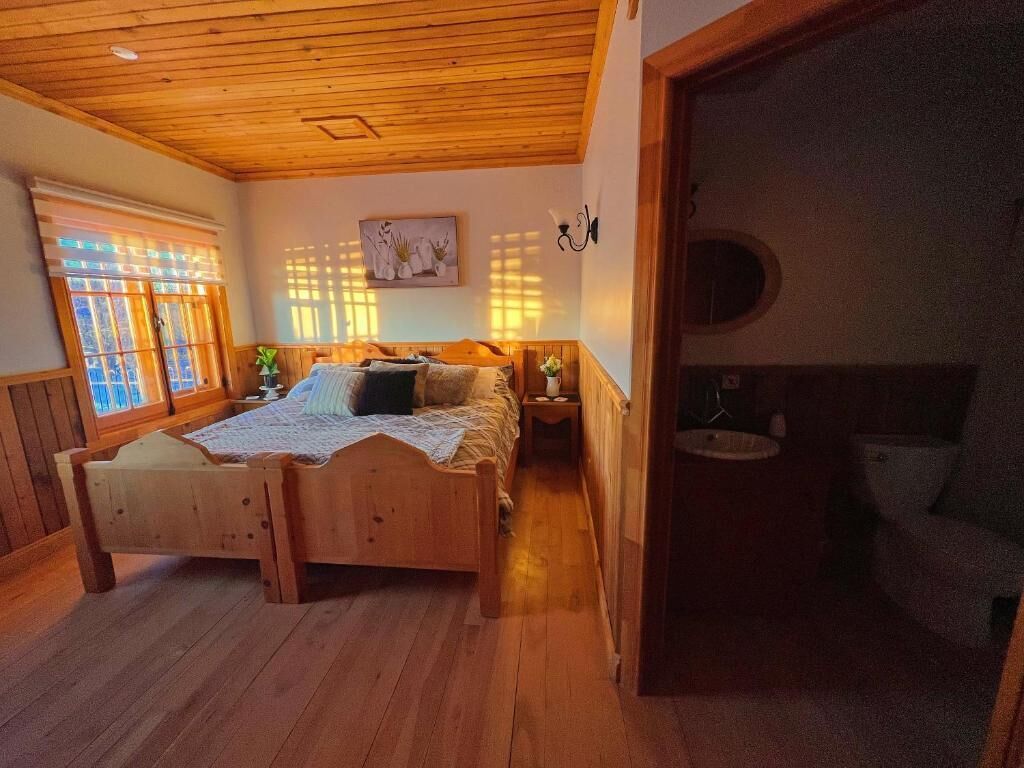 A wooden room with a bed, a nightstand, a window with blinds, and a door leading to another room.