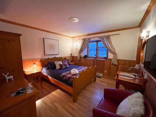 Un dormitorio con cama de madera, un escritorio con lámpara, una silla, una ventana con cortinas y un televisor.