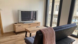Ein Wohnzimmer mit einer Couch, einem Fernseher auf einer Holzkommode, einem kleinen Tisch und großen Fenstern mit Blick auf Gebäude.
