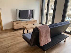 Ein Wohnzimmer mit einer Couch, einem Fernseher auf einer Holzkommode, einem kleinen Tisch und großen Fenstern mit Blick auf Gebäude.