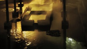 Eine nächtliche Szene mit Wasserspiel, Metallkonstruktion und einem Gebäude mit vergitterten Fenstern.
