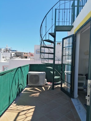 Una terraza en la azotea con una escalera de caracol, una barandilla de seguridad verde y una unidad de aire acondicionado.