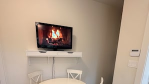 A dining area with a round table, four chairs, a flat-screen TV mounted on a white shelf, and a thermostat on the wall. - Peaceful Private Suite in Beautiful Home. 20 min to downtown (Burnaby)