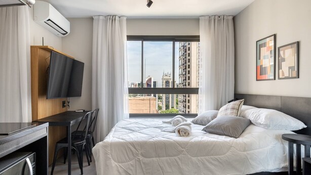 A hotel room with a bed, a desk, a TV, and a window with a city view. - Studio Novo Campo Belo Piscina Congonhas Moema (São Paulo)