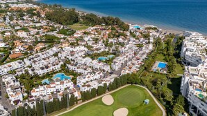 Vue aérienne d’un quartier résidentiel situé sur la côte, avec des bâtiments blancs, une plage et un terrain de golf.