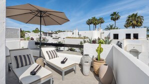 Une terrasse sur le toit avec des murs blancs, un parasol beige, deux chaises longues avec des coussins à rayures et des plantes en pot.