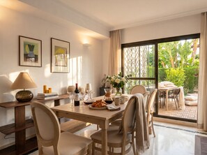 A dining area with a table set for six, a lamp, framed artwork, and a view of greenery through a large window. - Golf & Beach Villa beside Guadalmina Golf Club — sleeps 8, lagoon style pool (Marbella)