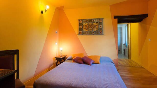 Quarto com cama, mesa de cabeceira, luminária na parede e porta para outro cômodo.