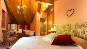 A bedroom with a wooden ceiling, a chandelier, a bed with floral bedding, a red pillow, a heart-shaped decoration on the wall, and a bathroom with a bathtub and toilet. - Bizet Suite: Passion and Charm in "Red" (Villair-Amerique)